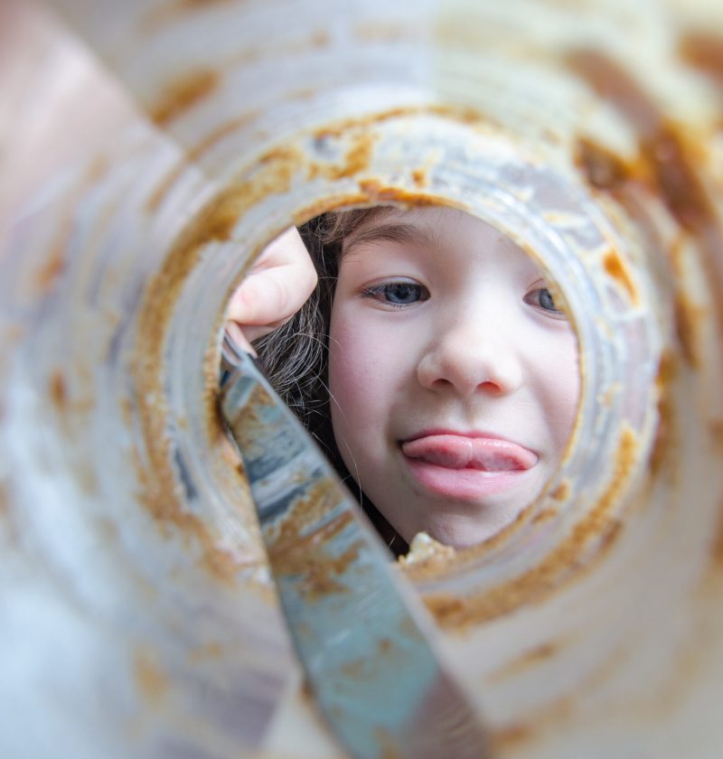 The face of a little gorl getting the last trace of peanut butter with a knife in the jar and sticking tongue out, seen from the bottom of the jar